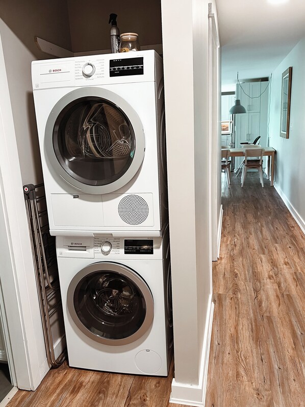 A stacked washer and dryer unit in a hallway with wooden flooring and a dining area in the background.