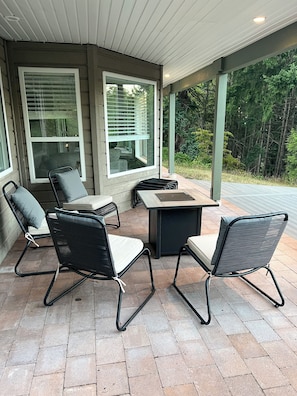 Eine Terrasse im Freien mit Tisch und vier Stühlen, großen Fenstern und Blick auf Bäume.