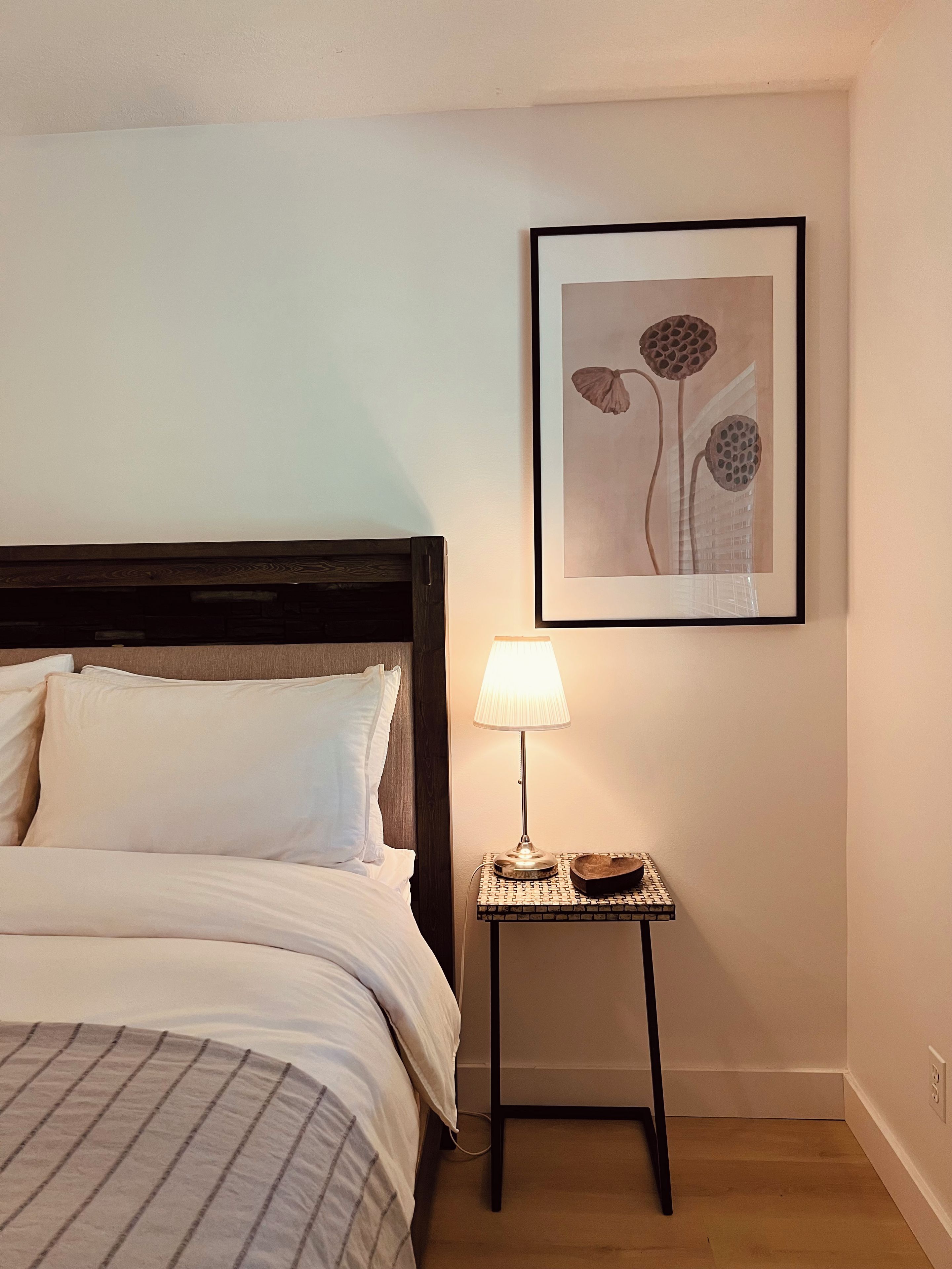 Un lit bien fait, avec une literie blanche, une petite table de chevet avec une lampe et, au mur, une œuvre d’art encadrée représentant des fleurs.
