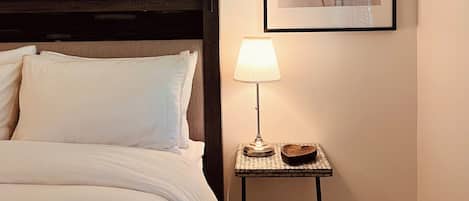 Un lit bien fait, avec une literie blanche, une petite table de chevet avec une lampe et, au mur, une œuvre d’art encadrée représentant des fleurs.