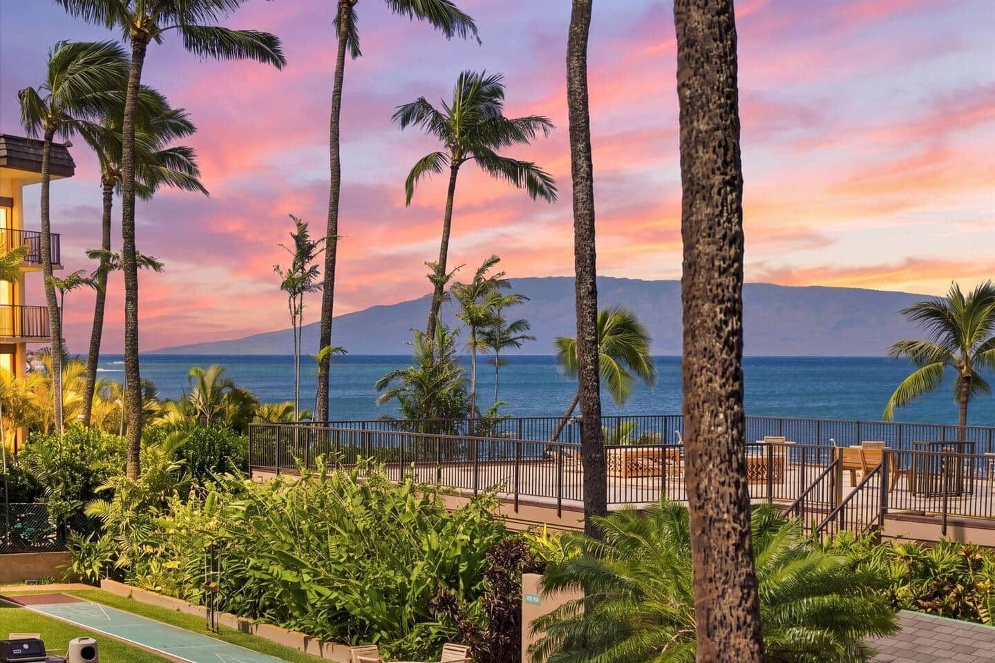 A resort with a view of the ocean and a mountain in the background, surrounded by palm trees and a fenced deck area.