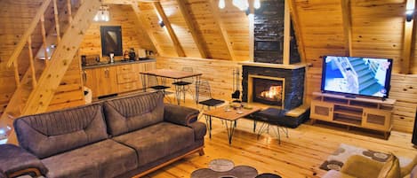 Et hyggeligt loftrum med træloft, sofa, spisebord, stole, pejs og fjernsyn.