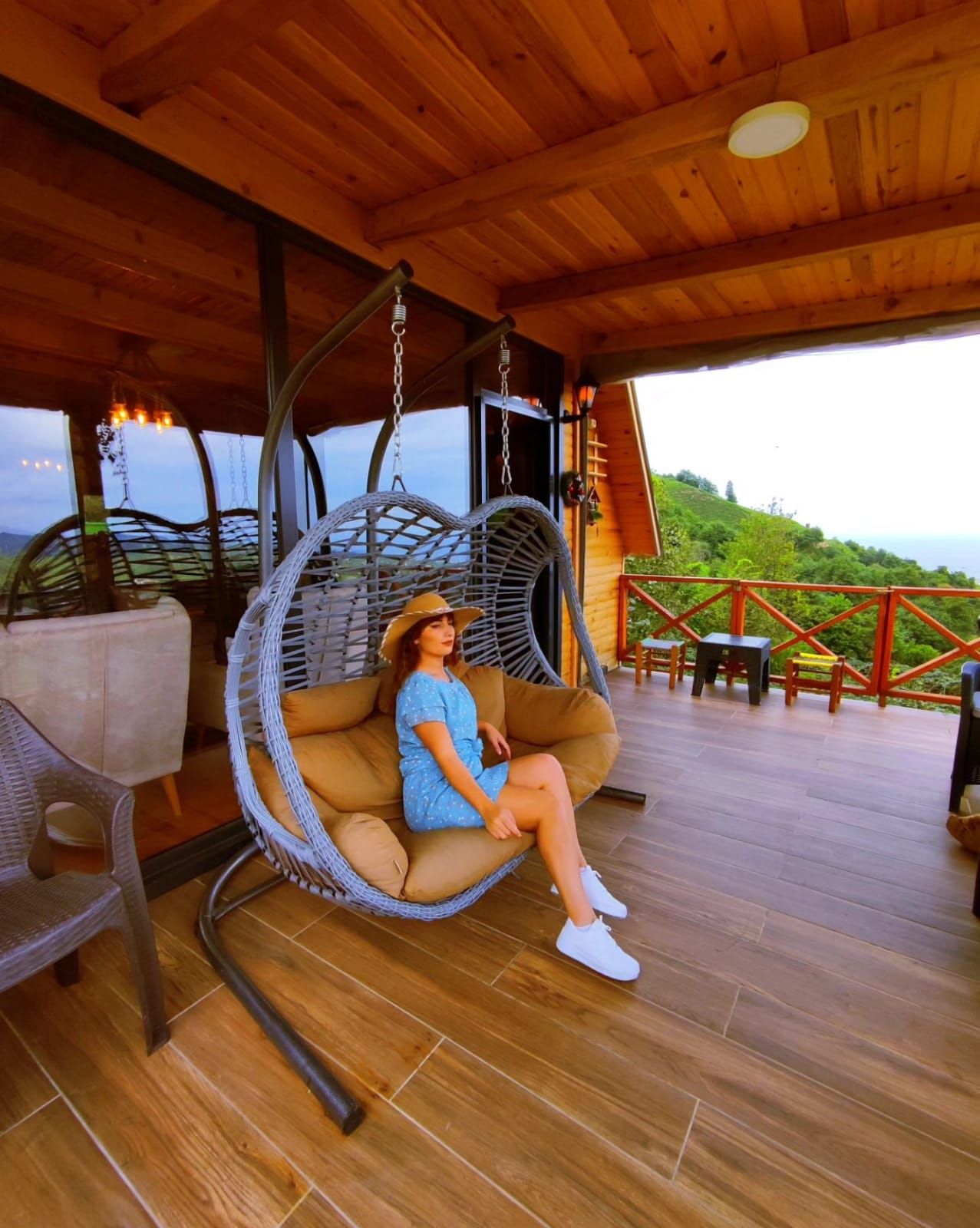 A person sitting on a hanging chair on a wooden deck with a view of greenery and hills.