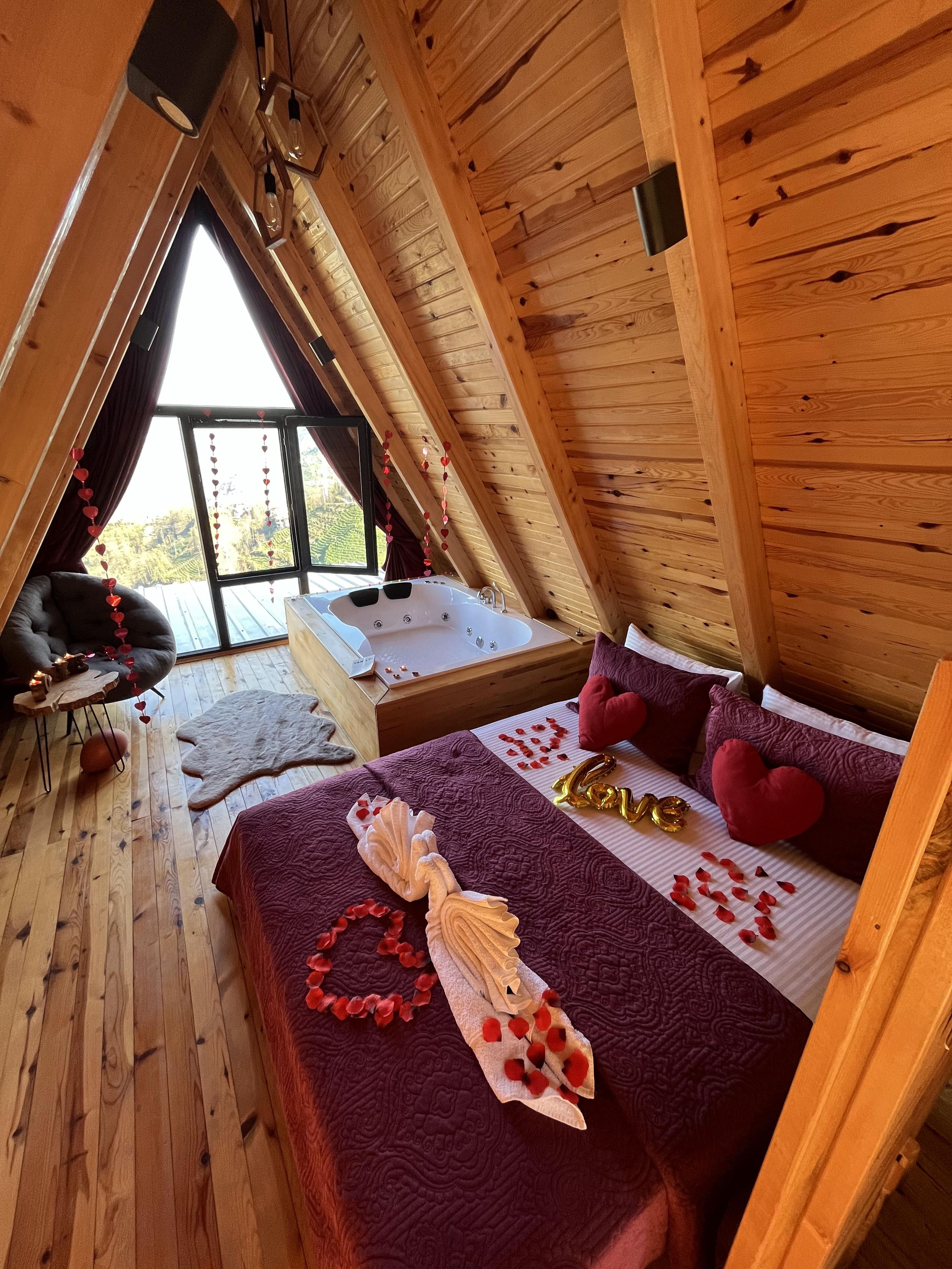 A cozy room with a sloped ceiling, a bed with a heart-shaped pillow, a small table with candles, and a view through a large window.
