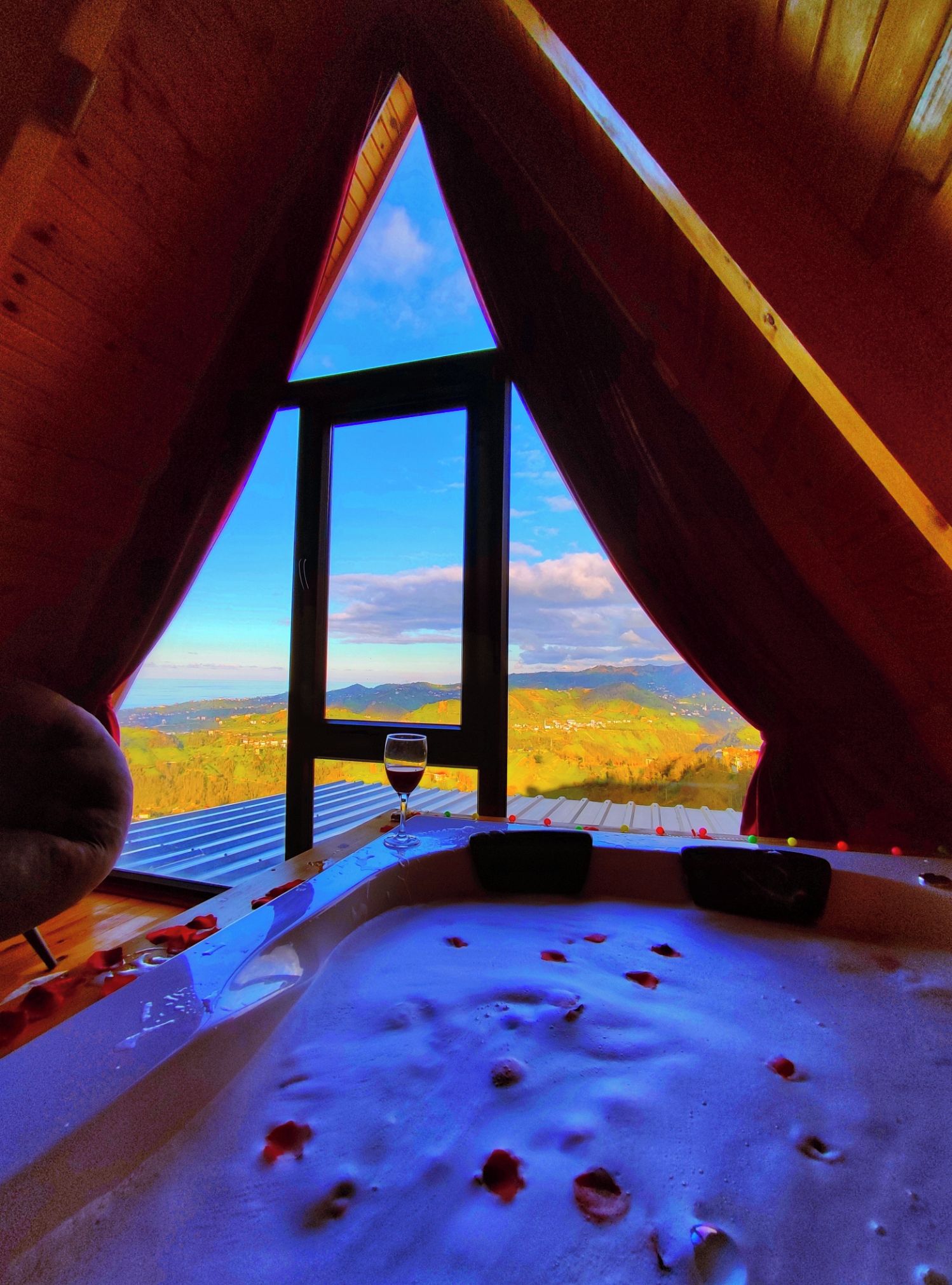 Vista interna de um quarto com uma grande janela, uma banheira de hidromassagem e um copo de vinho tinto.