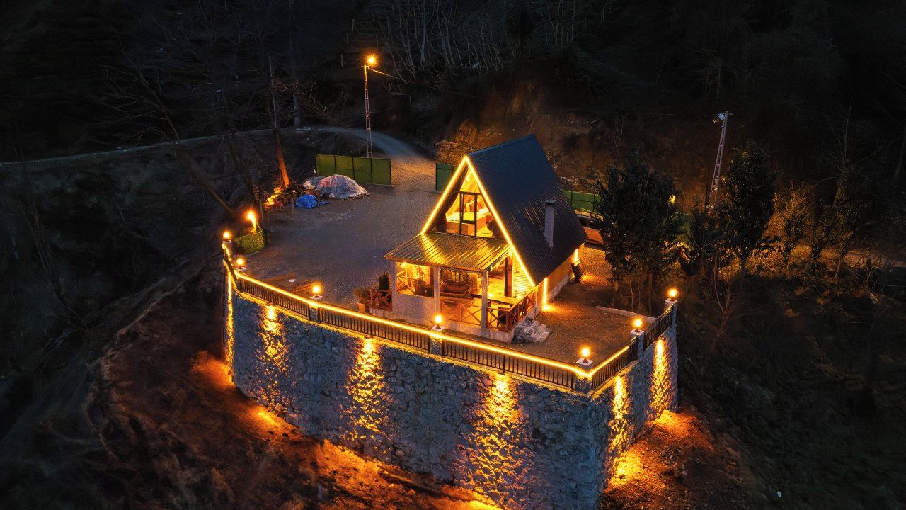 A house with a stone foundation, a fenced deck, and a lit interior.