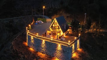 A house with a stone foundation, a fenced deck, and a lit interior.