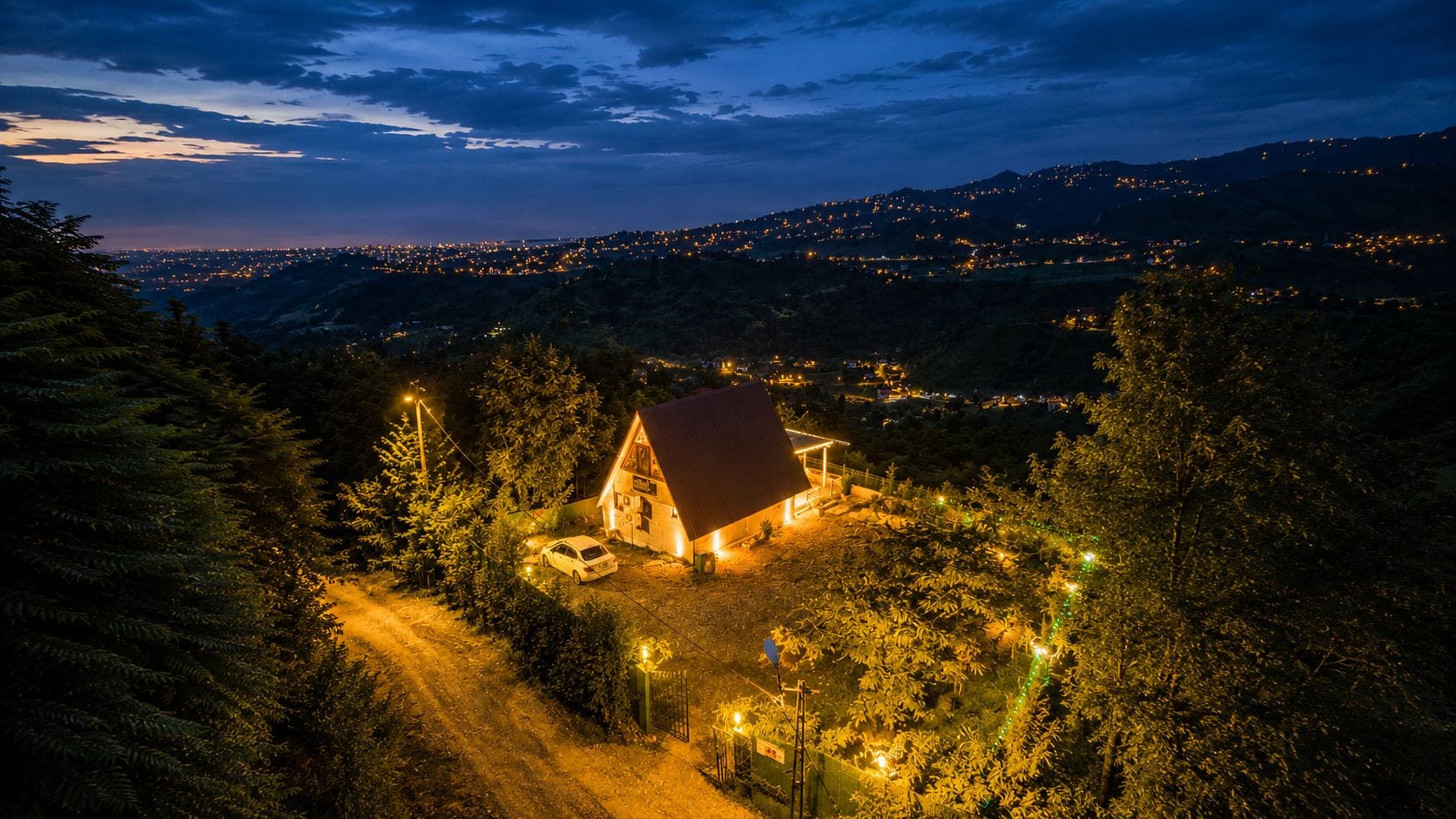 A house with a red roof surrounded by trees, located in a hilly area with distant city lights.
