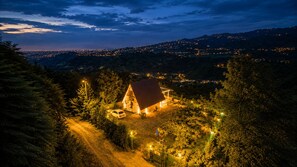A house with a red roof surrounded by trees, located in a hilly area with distant city lights.