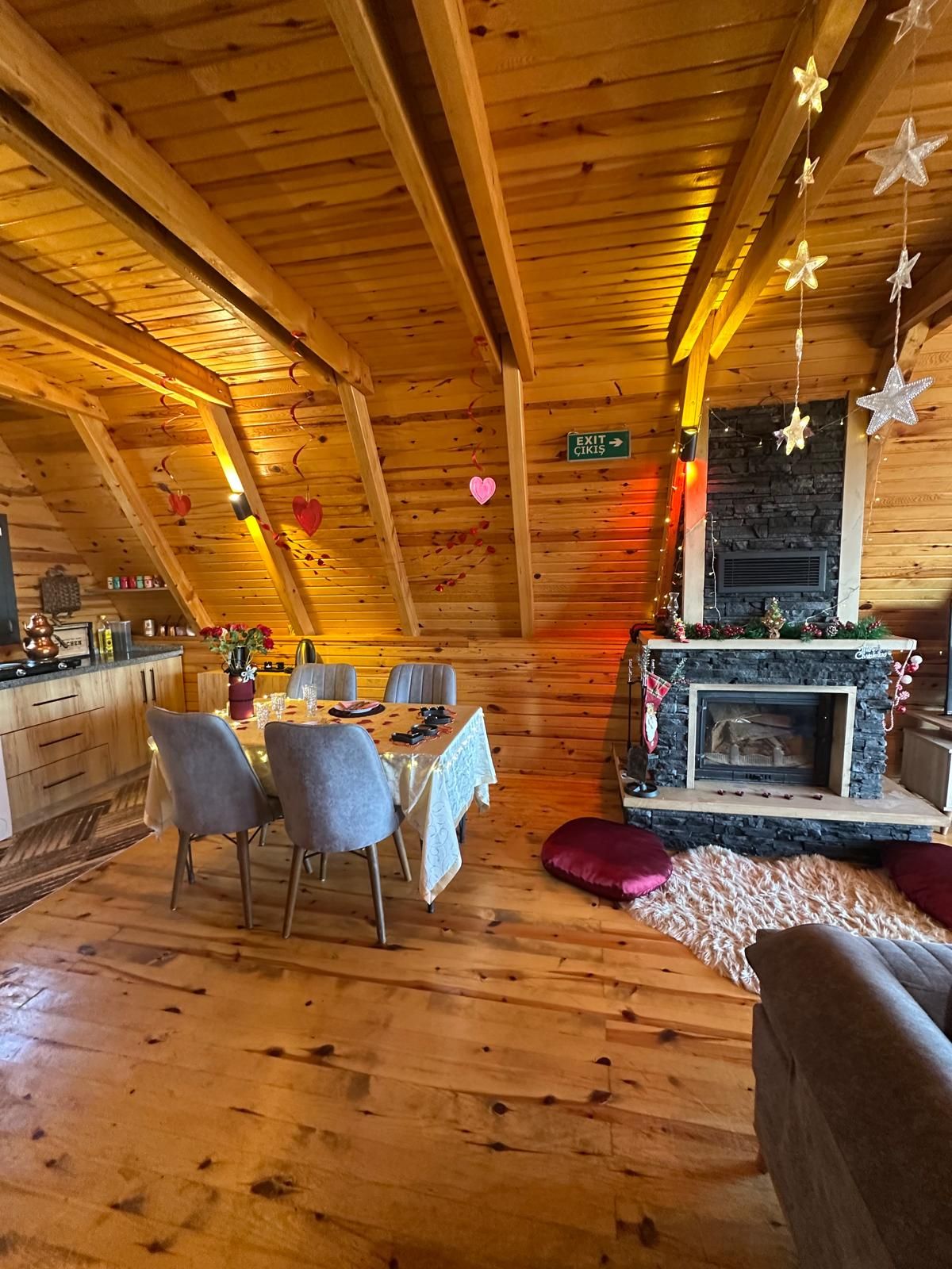 A cozy interior with a wooden ceiling, a dining area with a table and chairs, a fireplace, and a sign indicating an exit.