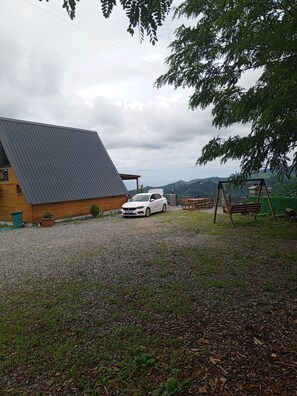 Une allée de gravier mène à un chalet en bois doté d’un toit en pente. Une voiture blanche est garée devant, et on aperçoit un portique à balançoire au fond.