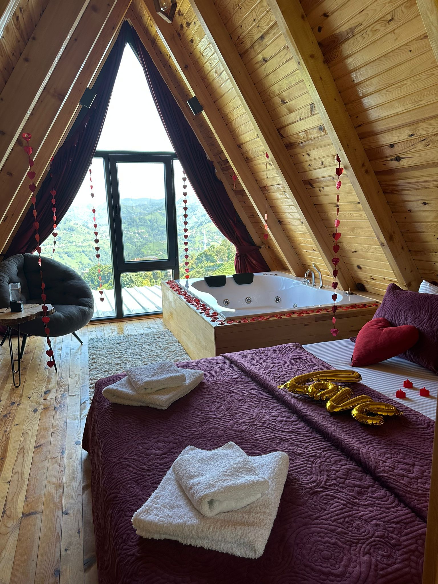 A cozy attic bedroom with a large window, a bathtub, a bed with towels, and a chair.