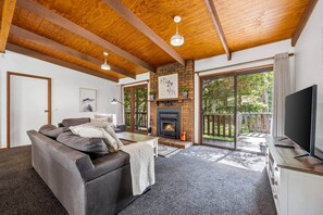A living room with a fireplace, a sofa, a TV, and a balcony with a view of greenery. - Tanglewood Cottage | Halls Gap Accommodation (Halls Gap)