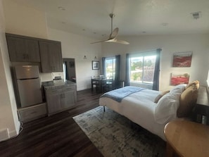 A compact hotel room with a kitchenette, a bed with a blue blanket, a dining area, and a large window with a view. - Serene Mountain & Temple View Retreat w/ Hot Tub Access (Riverton)