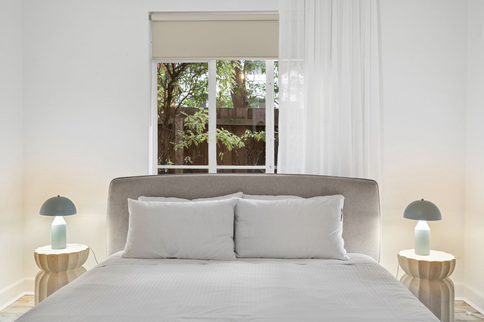 Ein Schlafzimmer mit einem Bett, zwei Nachttischlampen, einem Fenster mit Blick auf Grünflächen und einem weißen Vorhang.