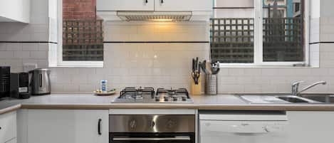 A modern kitchen with white cabinets, a gas stove, a dishwasher, and a window with a view of a brick wall and a lattice fence.