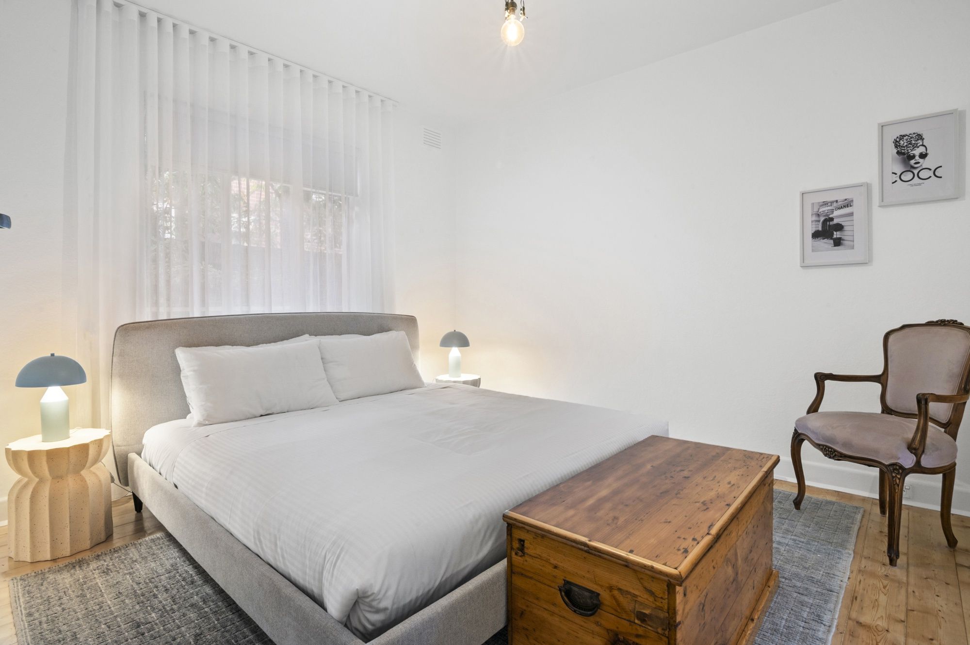 Une chambre à coucher comprenant un lit, des lampes de chevet, une commode en bois, une chaise et des œuvres d’art murales.