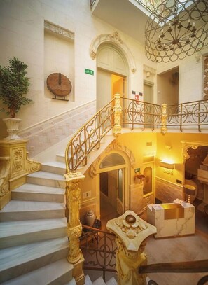 Una imponente escalera con barandillas decorativas y un candelabro, que conduce a un área del segundo piso con una recepción y una puerta que indica 'Exit'.