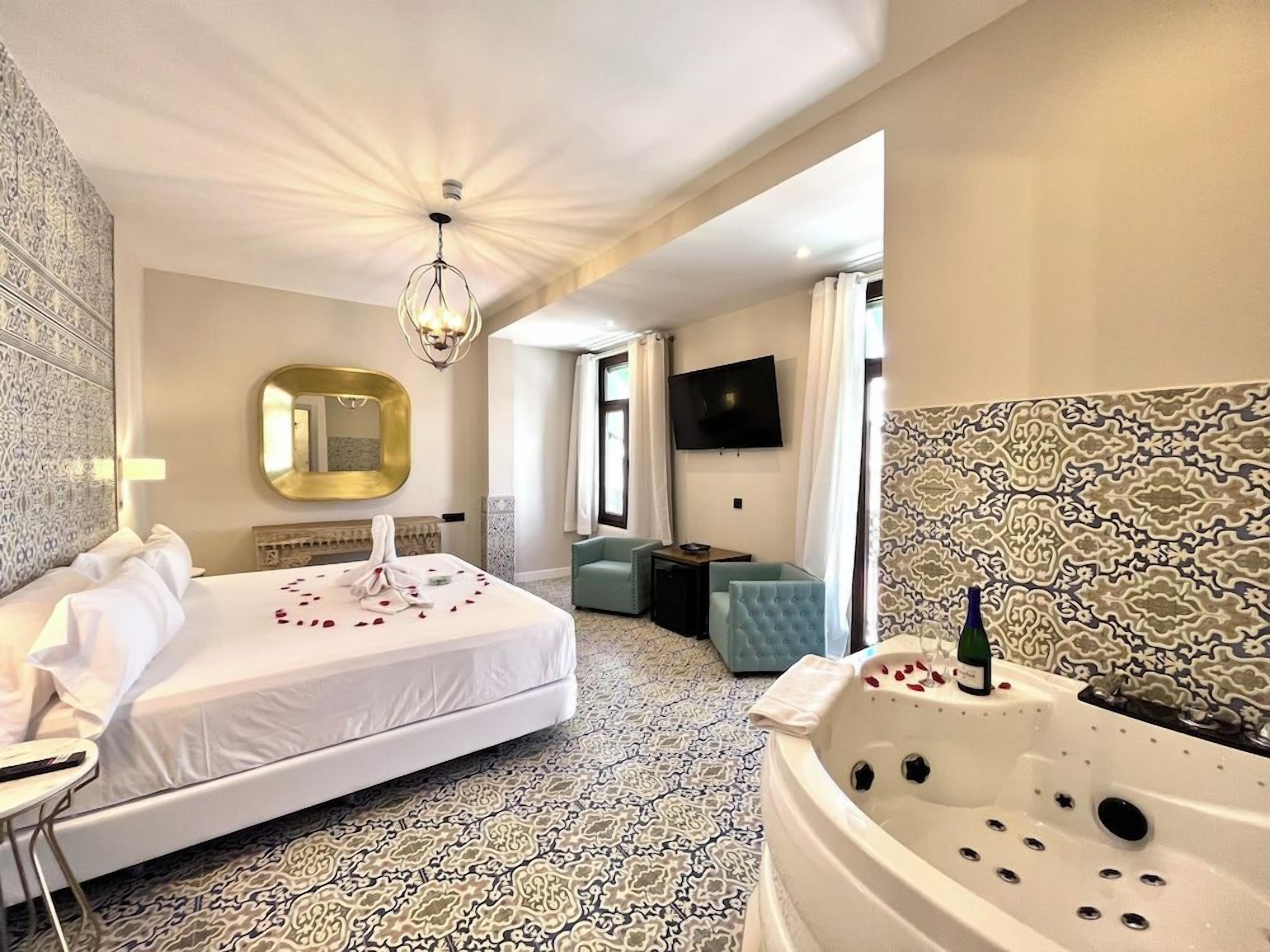 A hotel room with a large bed, a sitting area, a bathtub, and a television.