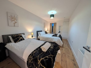 A hotel room with two beds, a nightstand, a lamp, a painting, a mirror, and a window with curtains. - Cozy Nest North Farm Rd Tunbridge Wells (Royal Tunbridge Wells)
