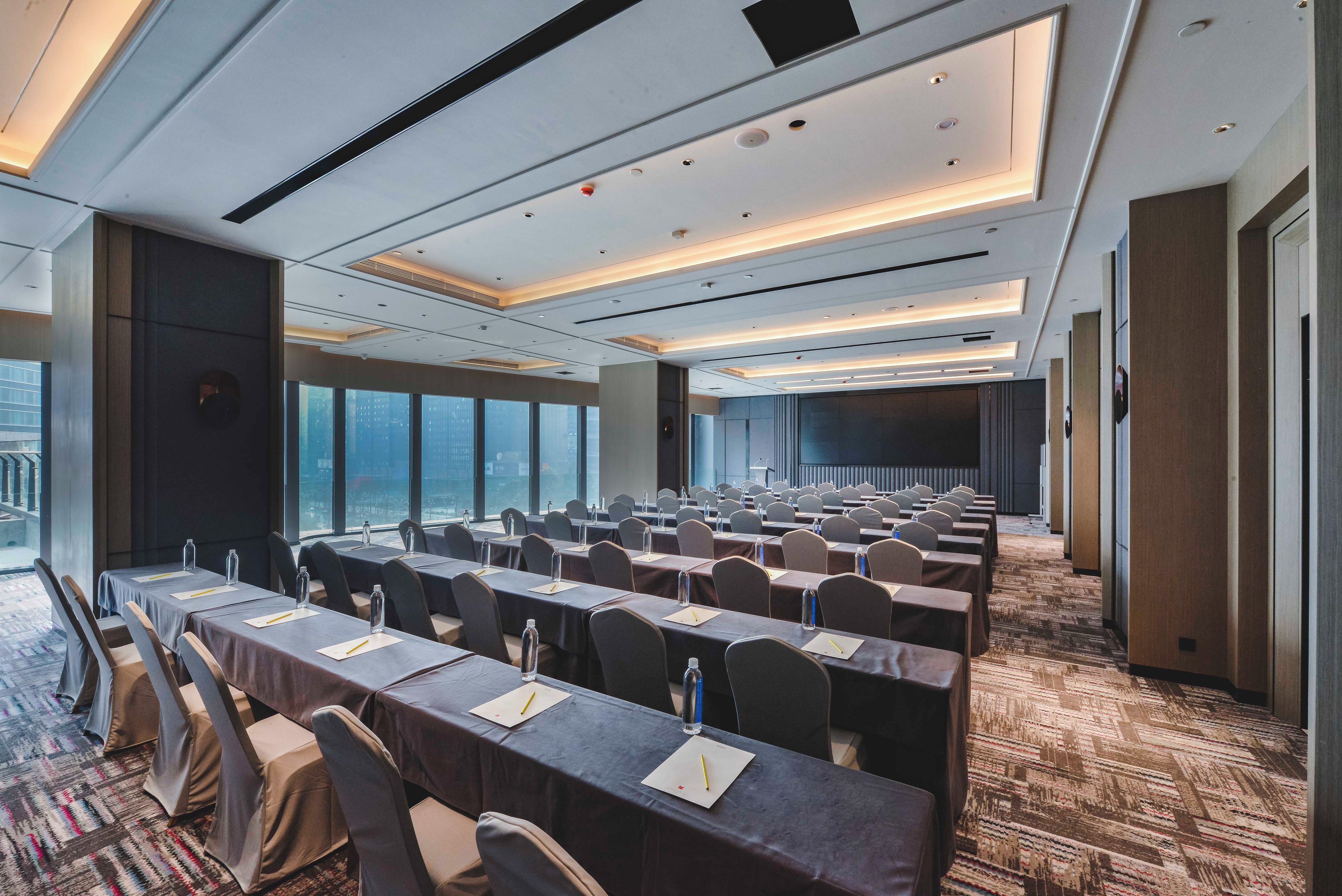 A conference room with rows of tables and chairs, large windows, and a carpeted floor.