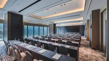 A conference room with rows of tables and chairs, large windows, and a carpeted floor.