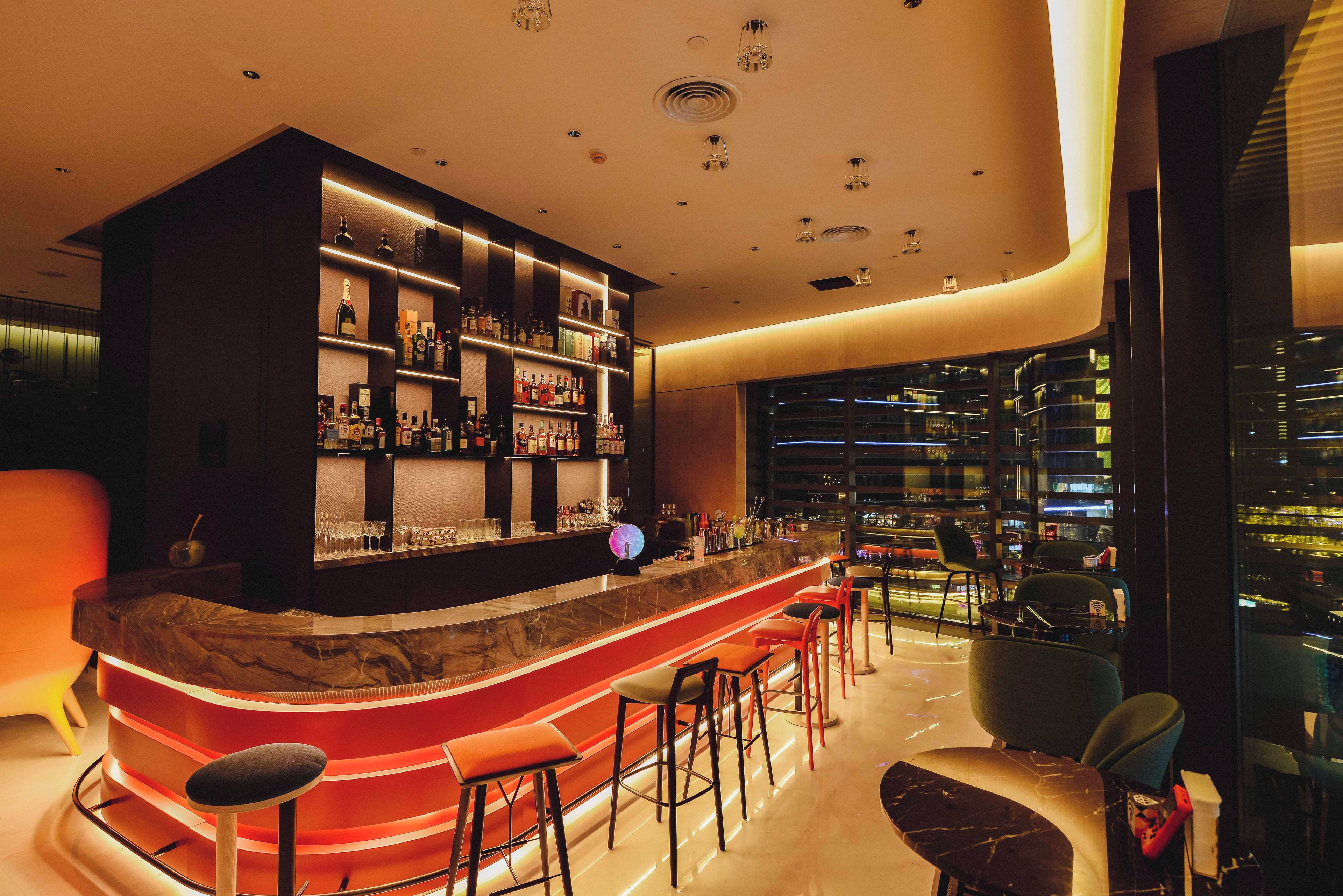 Zona de bar moderna con una barra curva, varios taburetes y una variedad de botellas y vasos en estanterías.