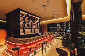 Zona de bar moderna con una barra curva, varios taburetes y una variedad de botellas y vasos en estanterías.