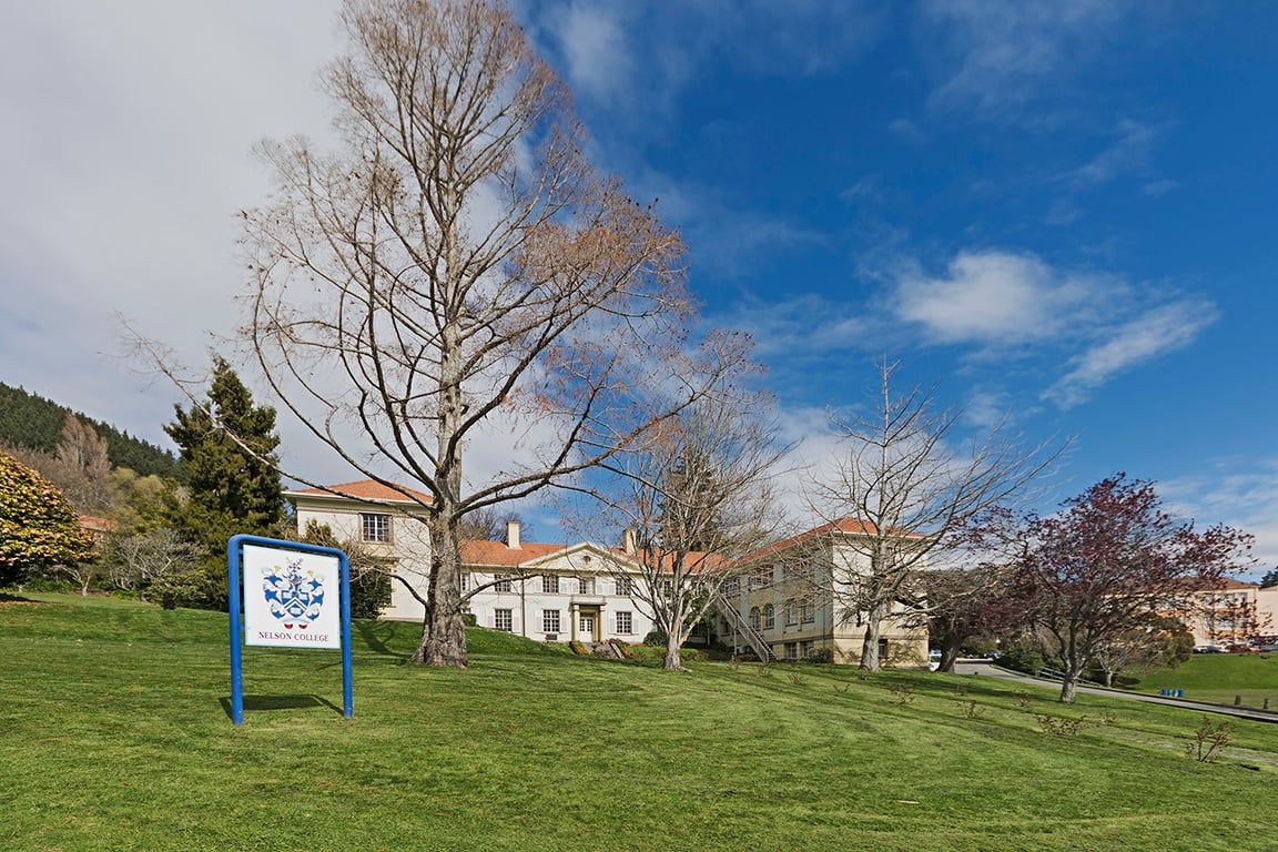Une zone herbeuse avec un panneau indiquant l’emplacement du Nelson College, un bâtiment doté d’un toit rouge et un ciel bleu dégagé.