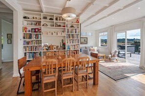 A spacious room with a wooden dining table, chairs, a bookshelf, and a view of the outdoors. - Sunset Paradise - Views, Hot Tub & Walk to Town (Nelson South)