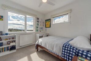 A bedroom with a bed, a desk, a chair, a radiator, a bookshelf, and two windows with curtains. - Sunset Paradise - Views, Hot Tub & Walk to Town (Nelson South)
