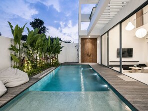 A modern outdoor pool area with a wooden deck, a seating area with cushions, and a large TV screen. - Casa Rio 3BR Enclosed Living & Pool (Keroboken Kelod)