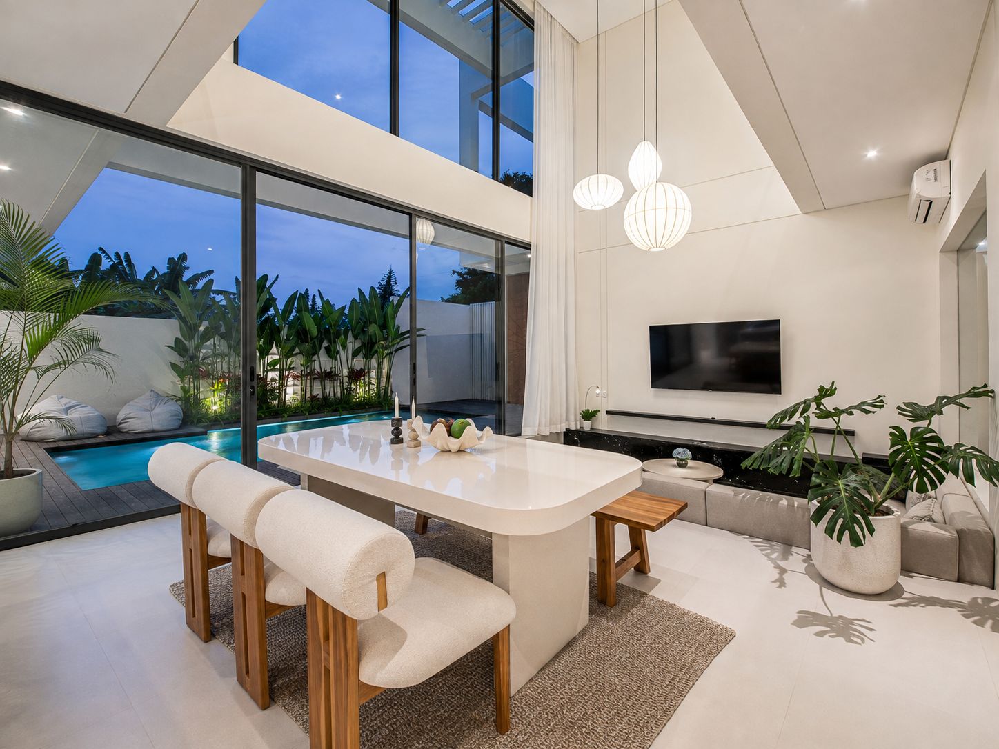 A modern living room with a large island table, seating, a flat-screen TV, and a view of a pool and tropical plants.