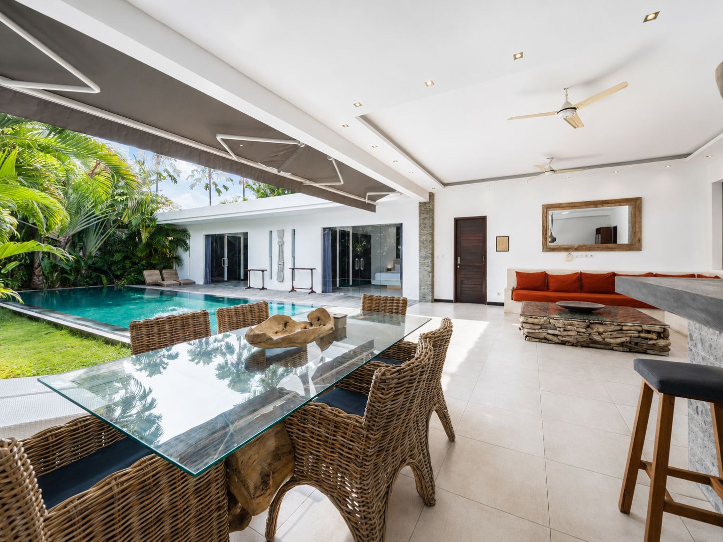 A modern interior with a glass dining table, wicker chairs, a stone accent piece, a sofa with orange cushions, and a pool outside.