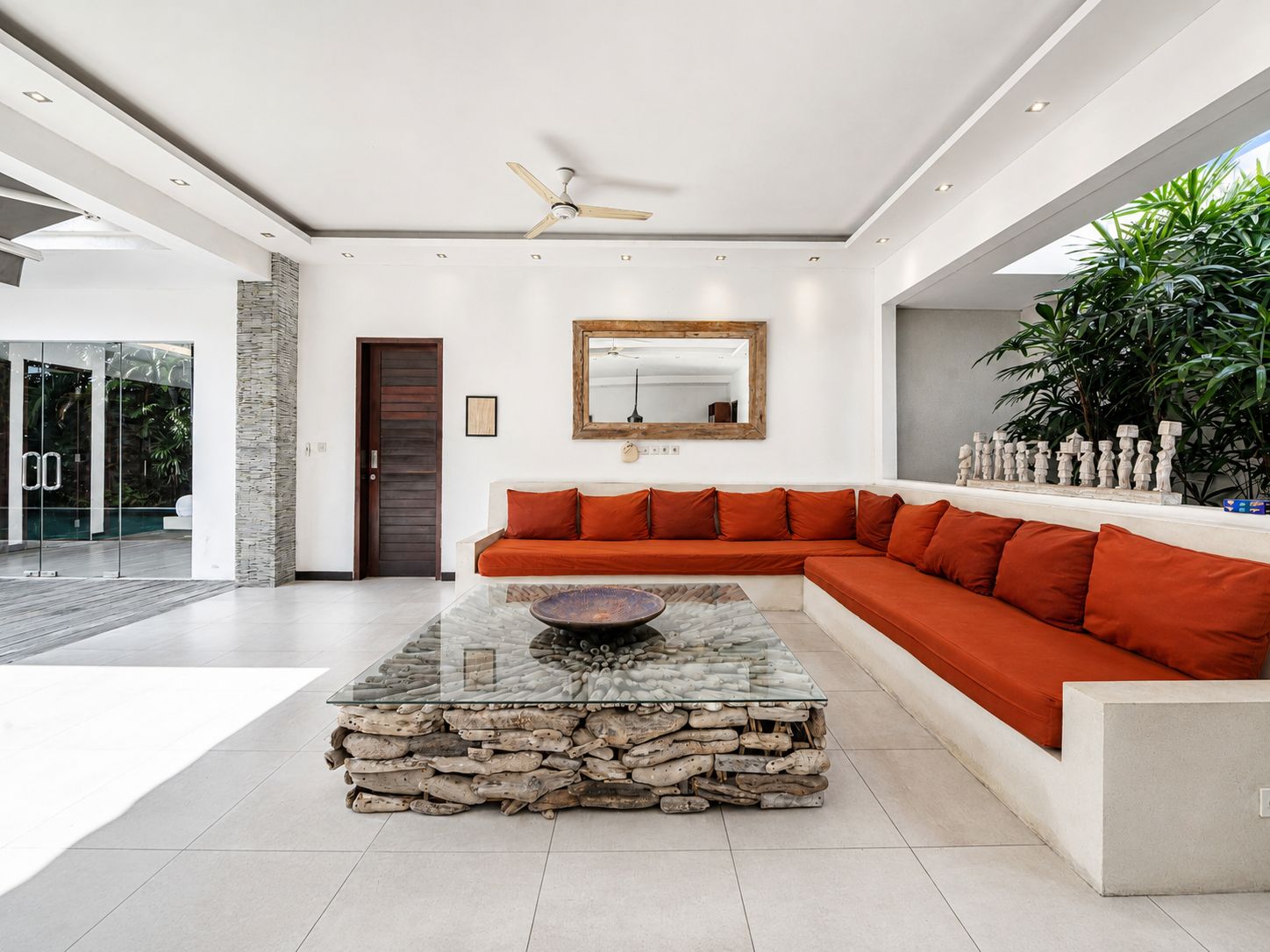 Una sala moderna con una mesa de centro de vidrio, una pared de acento de madera y un gran ventanal con plantas tropicales.