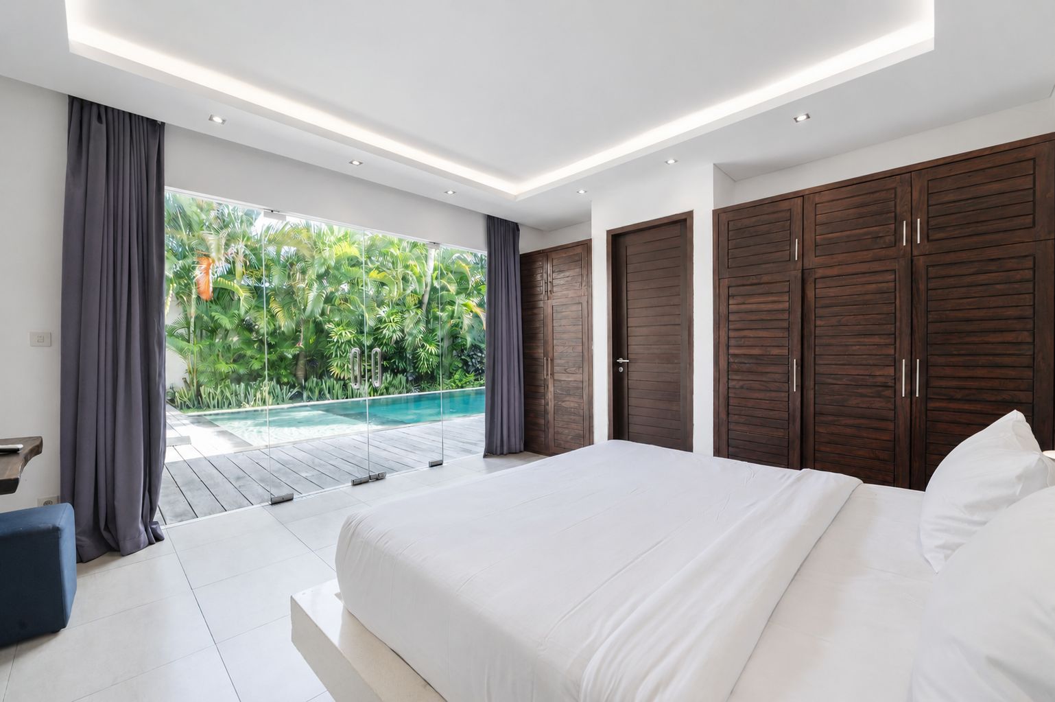 A modern bedroom with a large bed, wooden wardrobes, and a view of a swimming pool.