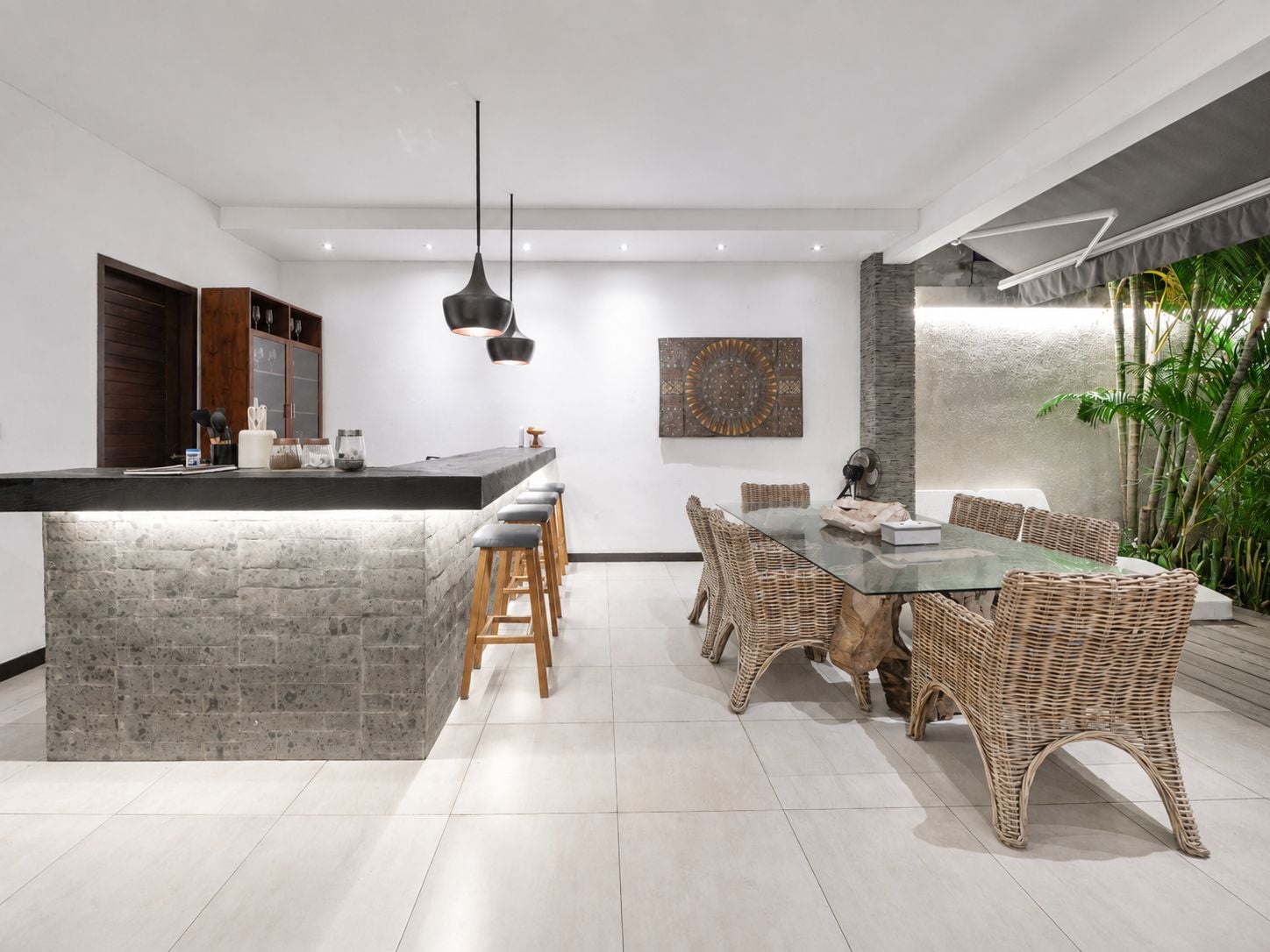 A modern kitchen with a central island, bar stools, a dining table with wicker chairs, and a pendant light.