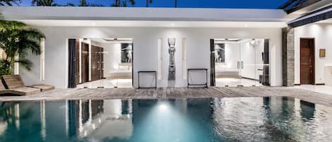 A modern poolside area with a clear pool, lounge chairs, and a glass-enclosed spa room.