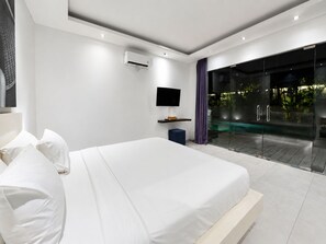 A modern bedroom with a large bed, a flat-screen TV, an air conditioning unit, and a glass door leading to an outdoor area. - Villa Juna Serene 3BR Hideaway Seminyak (Seminyak)