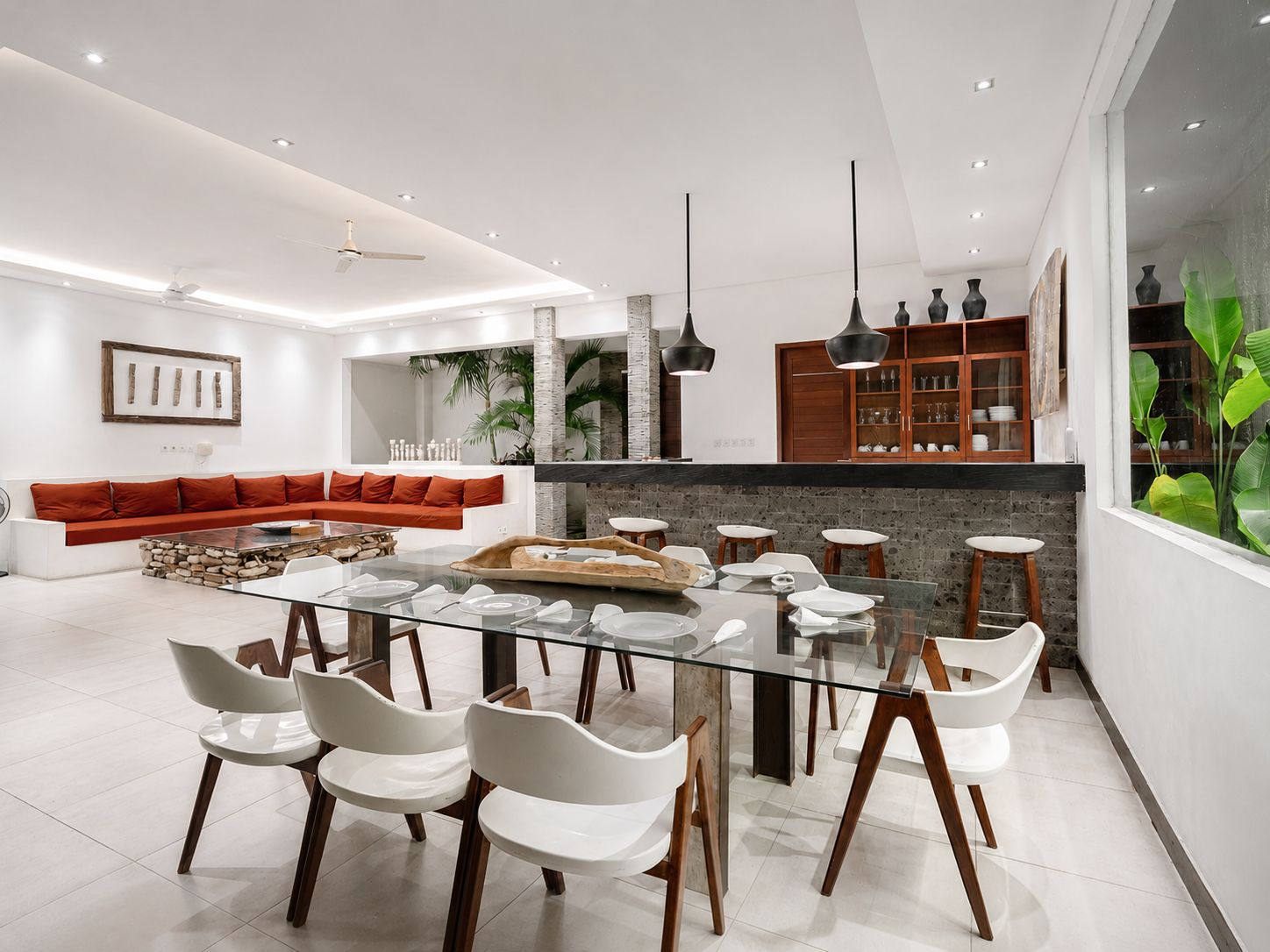 Un interior moderno con una mesa de comedor de vidrio rodeada de sillas blancas, un sofá rojo y una cocina con armarios de madera y luces colgantes.