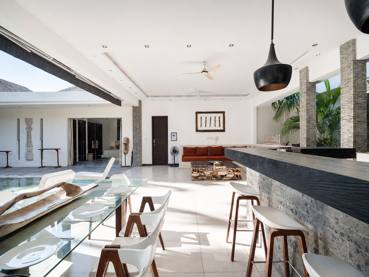 A modern interior with a glass dining table, seating, a ceiling fan, and a view of the outdoors.