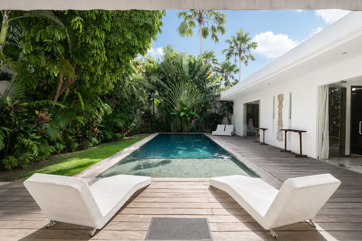 A modern outdoor pool area with a wooden deck, two white lounge chairs, and lush tropical vegetation.