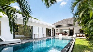 Ein moderner Poolbereich mit einem klaren, blauen Pool, umgeben von einem weißen Gebäude mit Glastüren und einer Terrasse mit Liegestühlen.