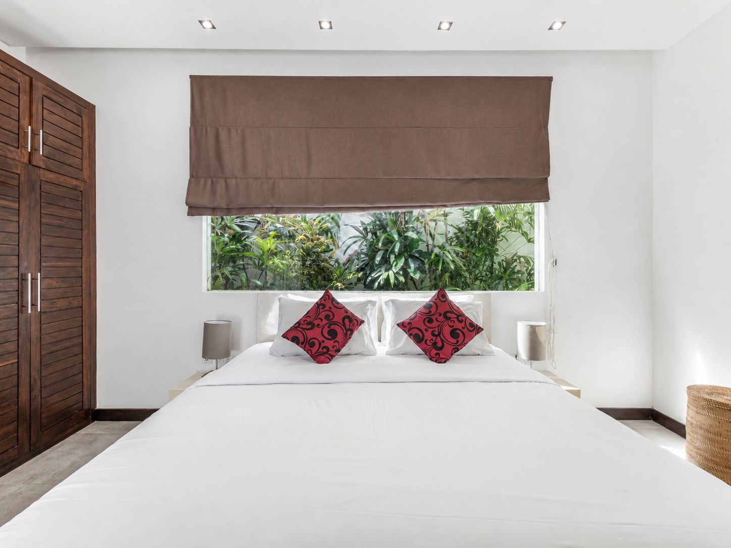 Une chambre avec un grand lit, deux oreillers, une fenêtre donnant sur un paysage verdoyant, une armoire en bois et un luminaire au plafond.