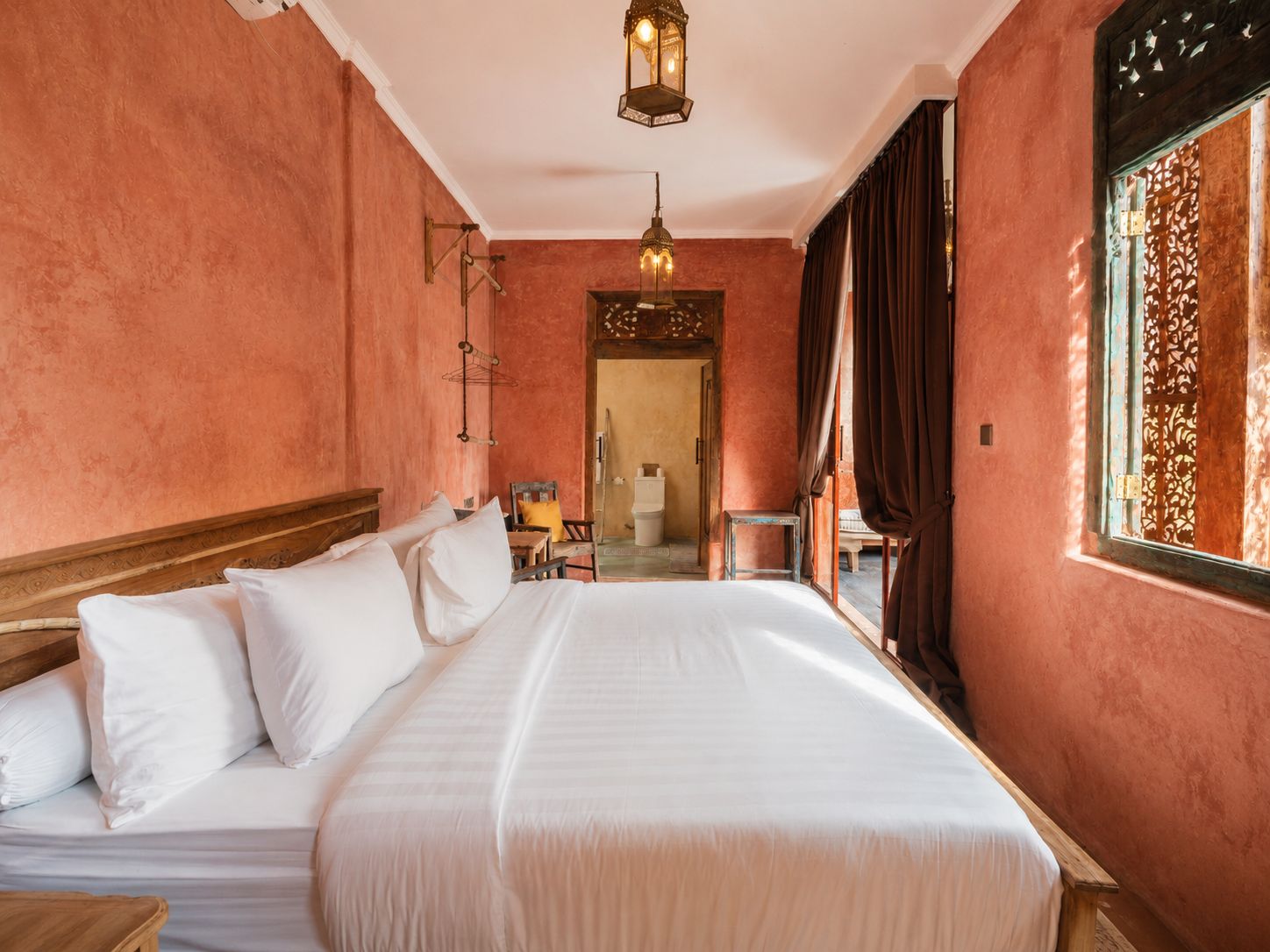 Een hotelkamer met een groot bed, een houten hoofdeinde, een hanglamp, een raam met gordijnen en uitzicht op de straat.
