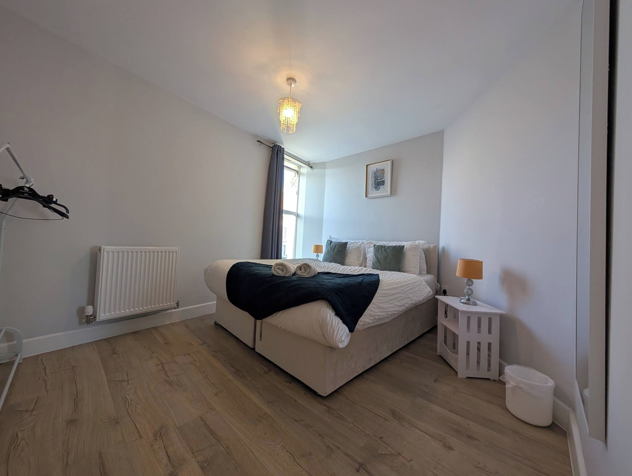 A bedroom with a bed, a bedside table with a lamp, a radiator, and a window with curtains.