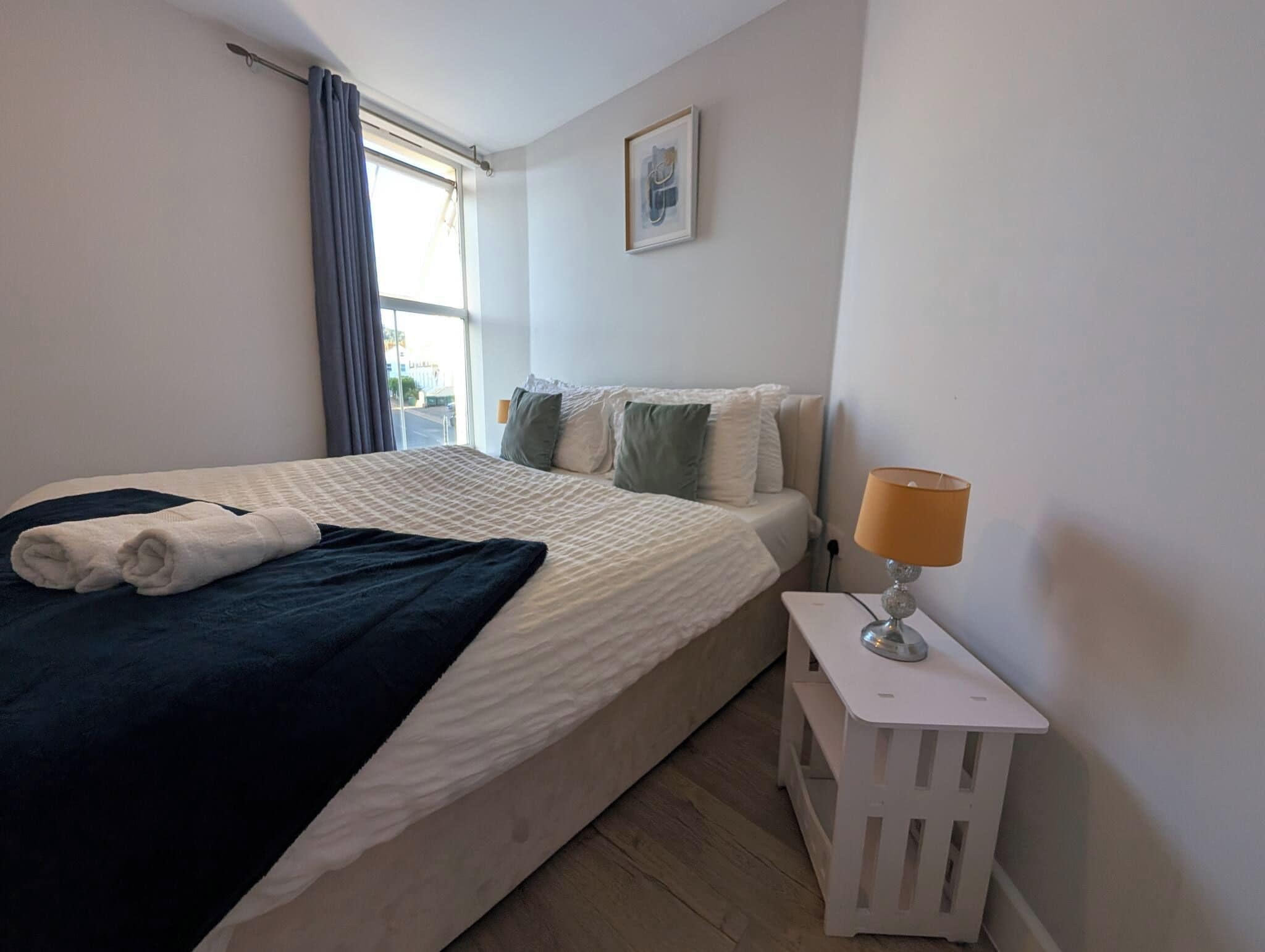 Et hotelværelse med en seng, et natbord med lampe, et vindue med gardiner og et billede på væggen.