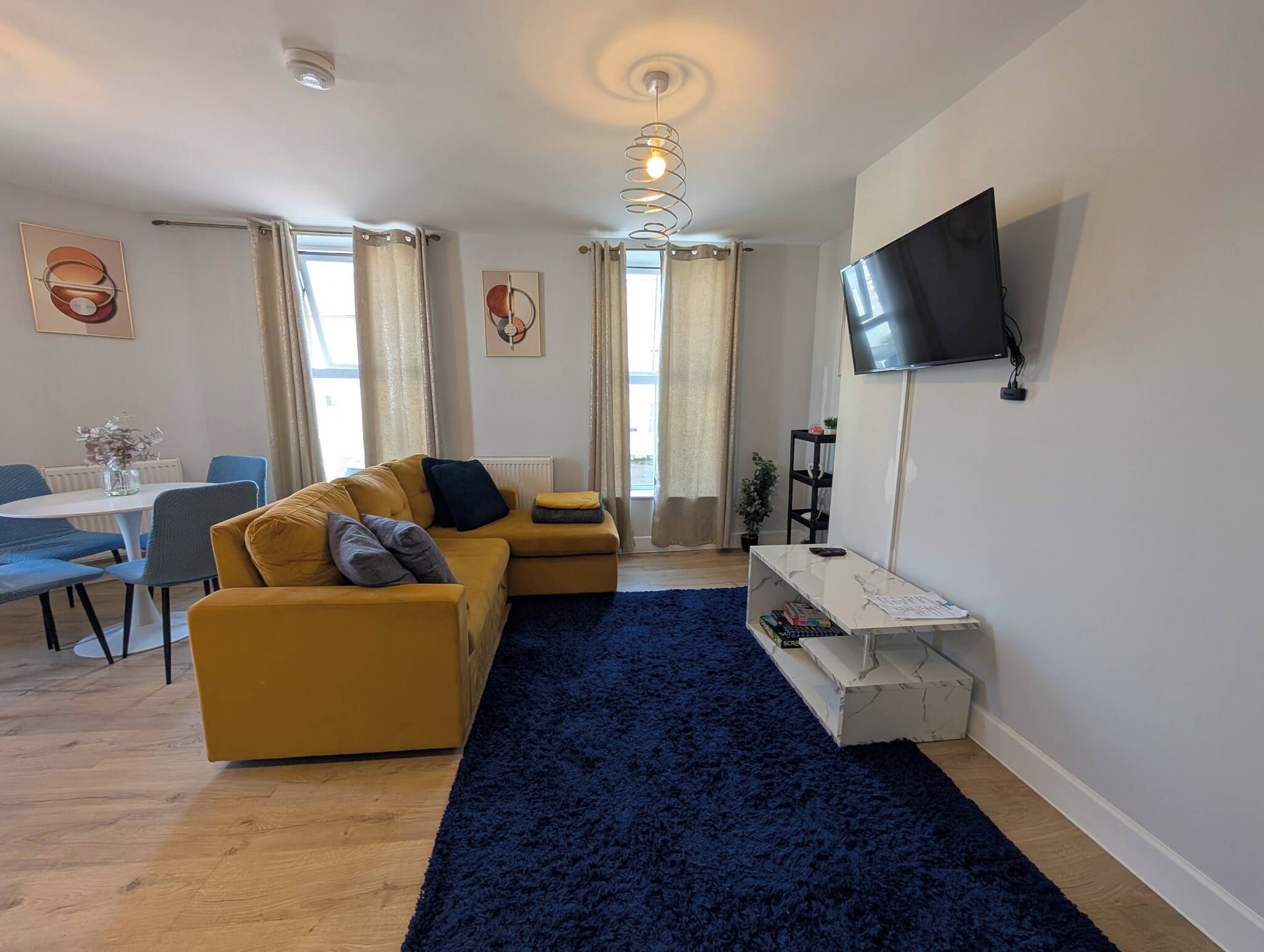 A modern living room with a sofa, a TV mounted on the wall, and a dining area with chairs and a table.