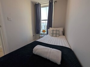 Una cama pequeña y ordenada con un edredón blanco y gris a cuadros, una manta azul oscuro y una toalla blanca. Hay una ventana con cortinas, una mesita de noche con una lámpara y una planta en maceta.