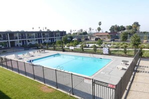 A rectangular swimming pool with a black metal fence, surrounded by a grassy area and a row of buildings in the background.